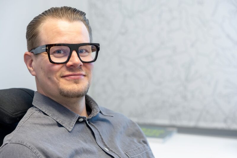 Mies harmaassa paidassa on lähikuvassa ja istuu.