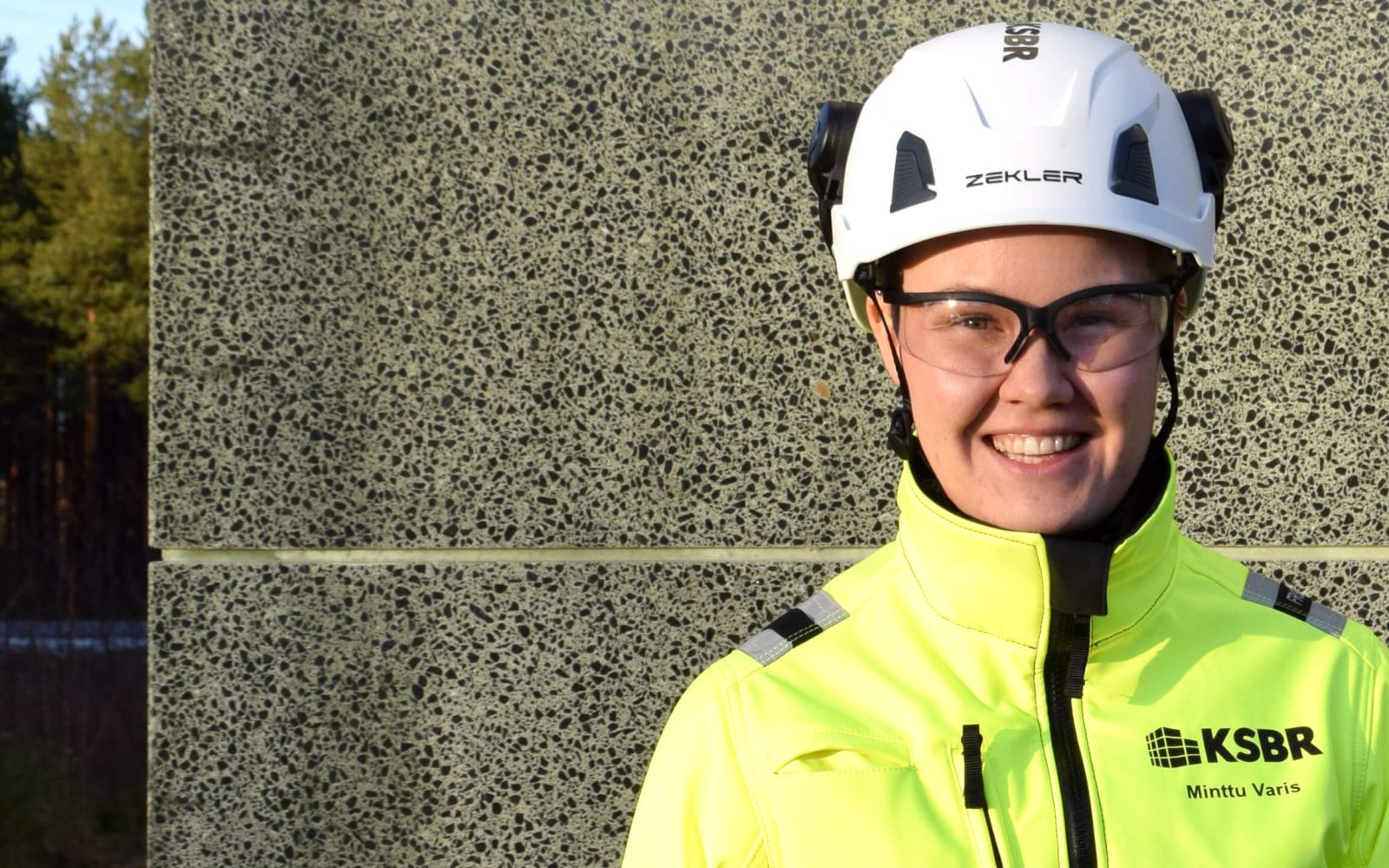 Hymyilevä rakennustyöntekijä seisoo betoniperustuksen edessä.