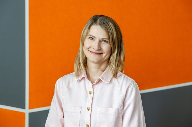 Nainen vaaleassa paitapuserossa seisoo oranssiharmaan seinän edessä ja hymyilee.