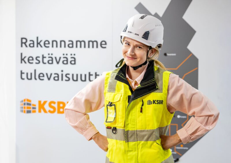 Työturvallisuuspäällikkö seisoo harmaan seinän edessä yllään kypärä ja huomioliivi.