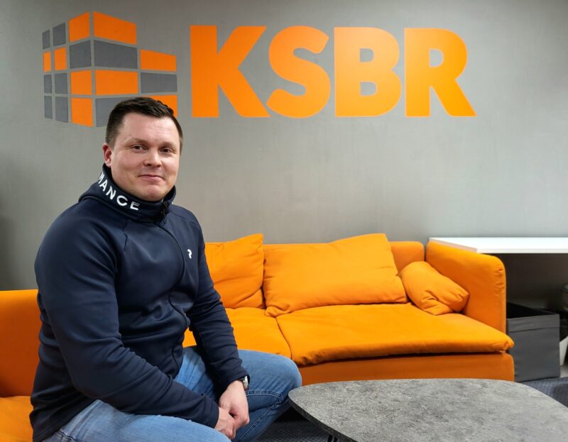 Toimitusjohtaja Marko istuu oranssilla sohvalla seinällä olevan KSBR:n logon edessä.