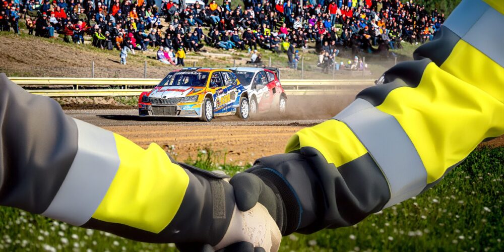 Kaksi kättä kättelee etualalla, taustalla kulkee oranssi ralliauto.