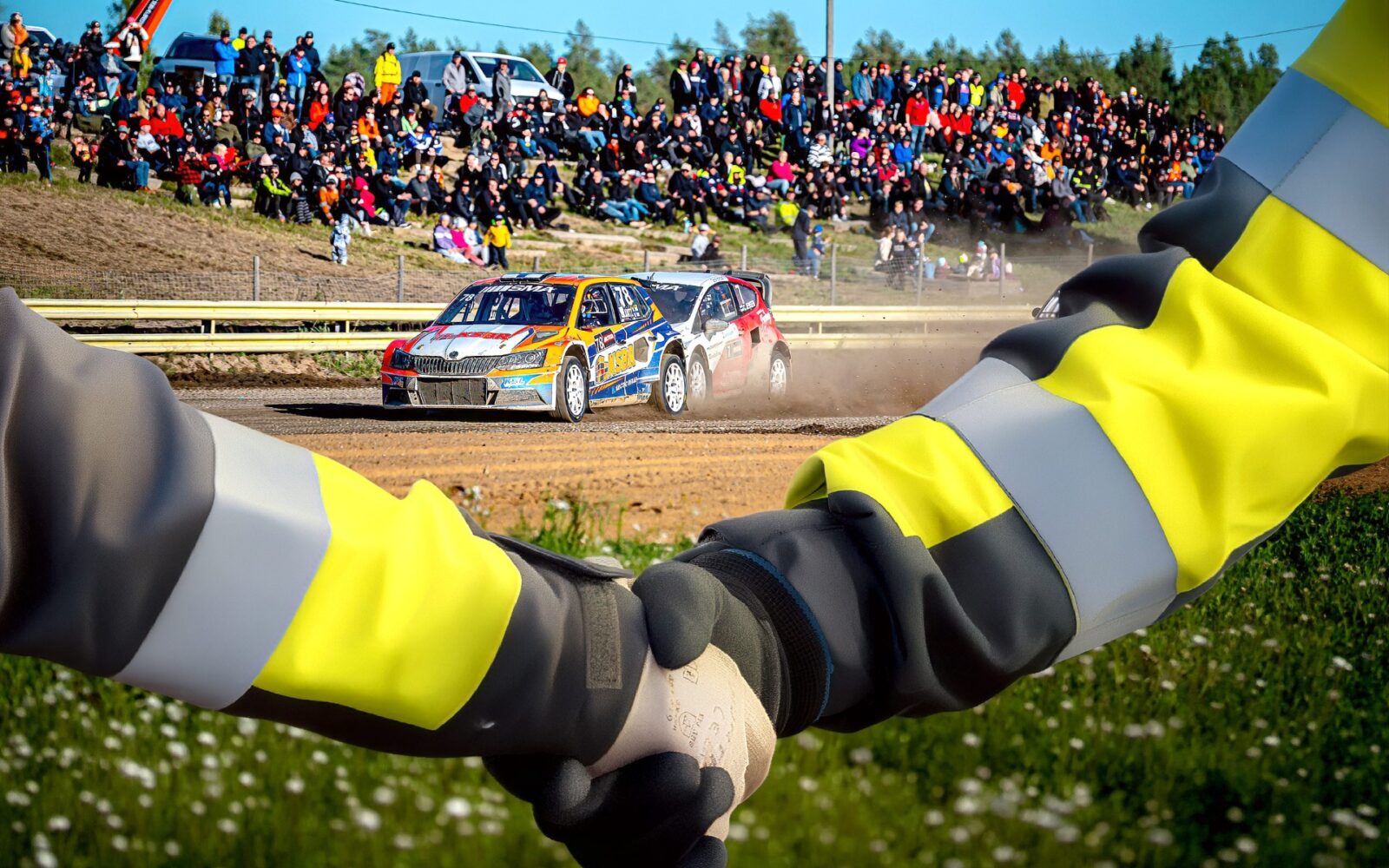 Kaksi kättä kättelee etualalla, taustalla kulkee oranssi ralliauto.