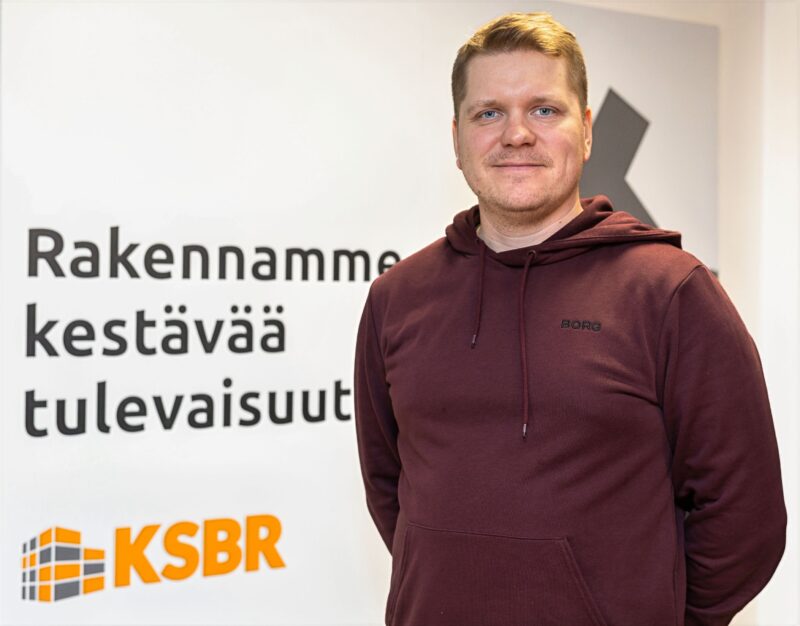 Punapaitainen mies seisoo KSBR-logon edessä hymyillen kädet selän takana.