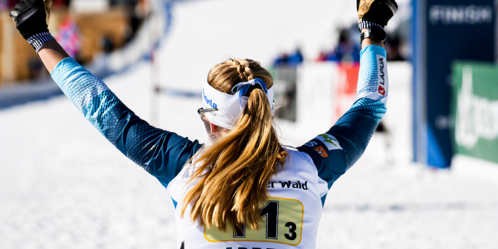 Vaalea hiihtourheilijanainen seisoo selin ja tuulettaa kädet ylhäällä mitalia.