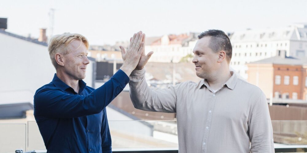 Kaksi yritysjohtajaa laittaa kädet yhteen taustallaan kaupunkinäkymä.