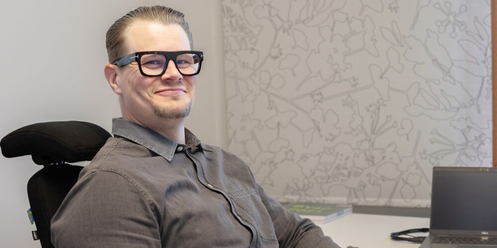 Mies harmaassa paidassa istuu tietokoneen ääressä.
