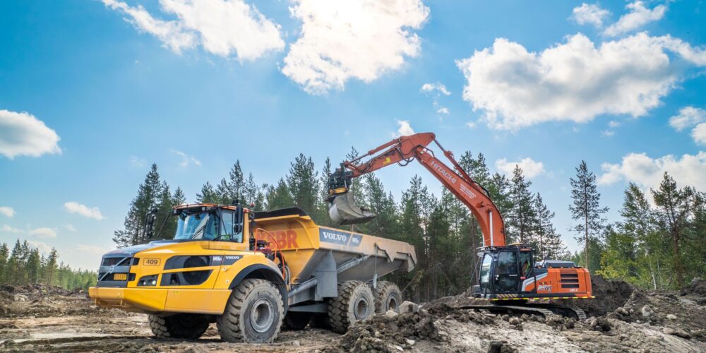 Maansiirtokone kaivaa maata aurinkoisessa säässä metsäisellä alueella.