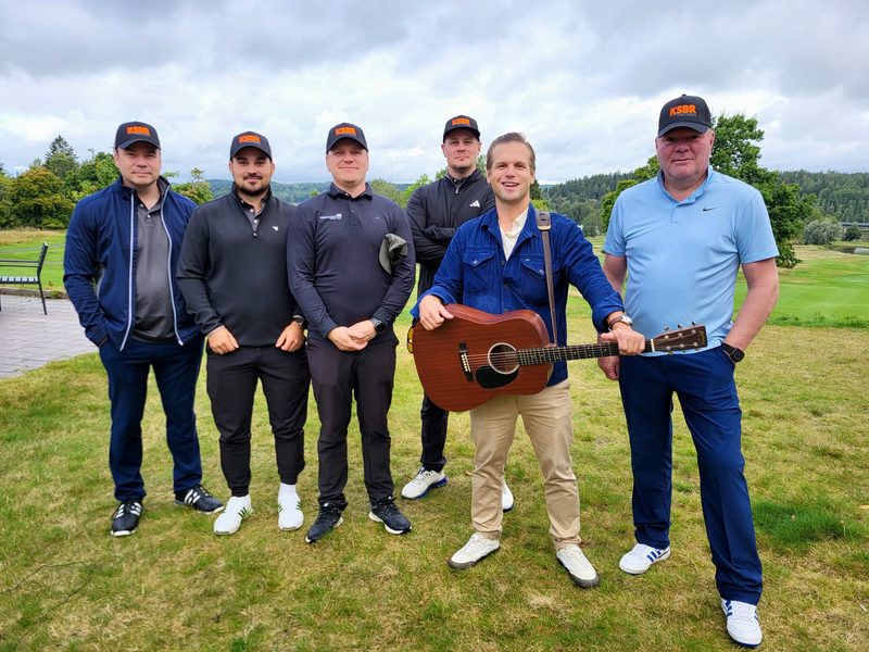 Työporukkaa seisoo golfkentällä kitaristin kanssa.